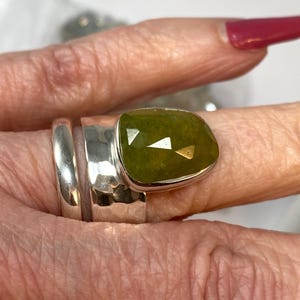 May include: A silver ring featuring a large, faceted, olive-green gemstone set in a bezel. The ring band consists of a smooth, thin silver wire paired with a wider, hammered silver band that supports the prominent, irregularly shaped stone.
