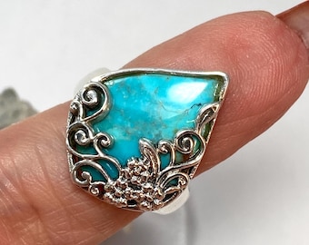 May include: A silver ring featuring a teardrop-shaped turquoise stone set within an ornate metal frame. The silver setting displays intricate scrollwork and small, textured floral-like details that wrap around the base of the bright blue stone.