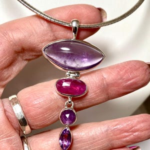 May include: A silver necklace with a pendant featuring a large purple amethyst gemstone, a smaller pink ruby gemstone, and two smaller purple amethyst gemstones.