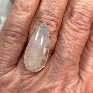 May include: A silver ring featuring a large, teardrop-shaped, translucent gemstone. The gemstone has a pale pink hue with internal veining. The ring is set on a silver band and is displayed on a hand.