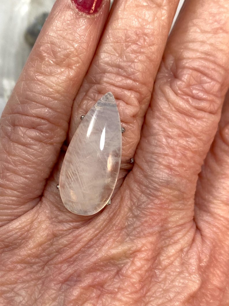 May include: A silver ring featuring a large, teardrop-shaped, translucent gemstone. The gemstone has a pale pink hue with internal veining. The ring is set on a silver band and is displayed on a hand.