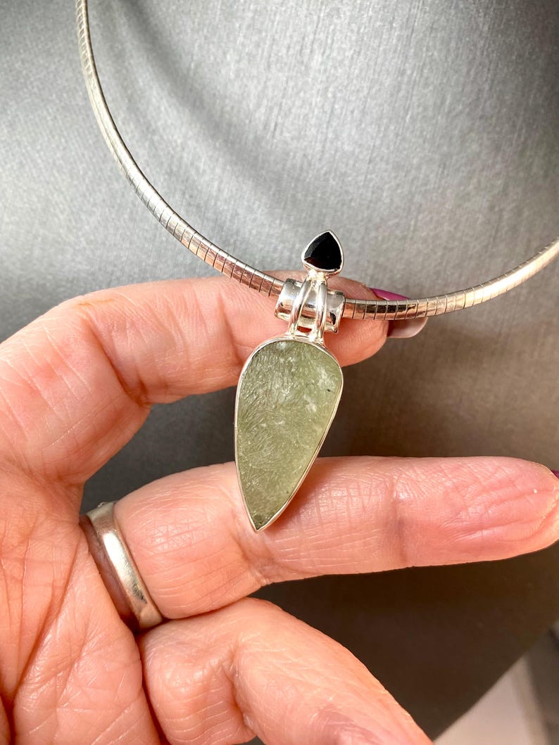 May include: A silver chain necklace with a pendant featuring a light green stone and a black gemstone. The pendant is shaped like a teardrop.