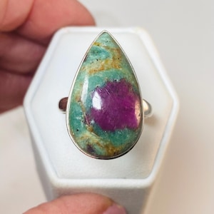 Puede incluir: Un anillo de plata con una piedra preciosa grande en forma de lágrima. La piedra presenta una vibrante mezcla de colores, con un magenta profundo en el centro, rodeado de verde y dorado. El anillo se muestra sobre una base blanca.