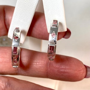 May include: A pair of silver hoop earrings with pink gemstones. The earrings are set with rectangular and square shaped stones.
