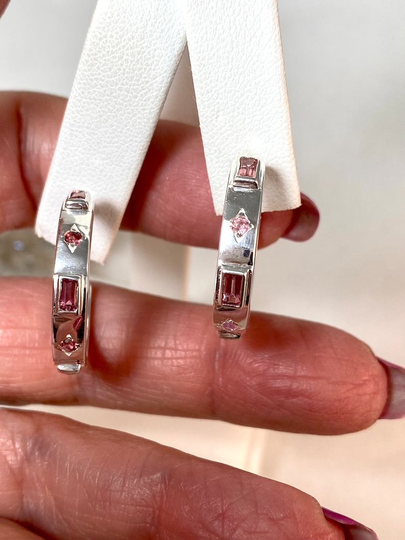 May include: A pair of silver hoop earrings with pink gemstones. The earrings are set with rectangular and square shaped stones.