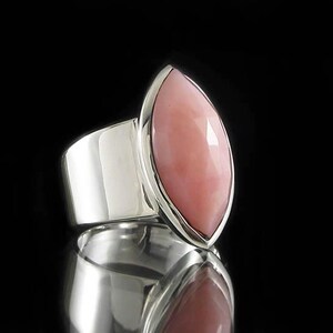 Pode incluir: Um anel de prata com uma grande pedra de opala rosa em forma de marquesa. O anel tem uma banda larga e um acabamento liso e polido.
