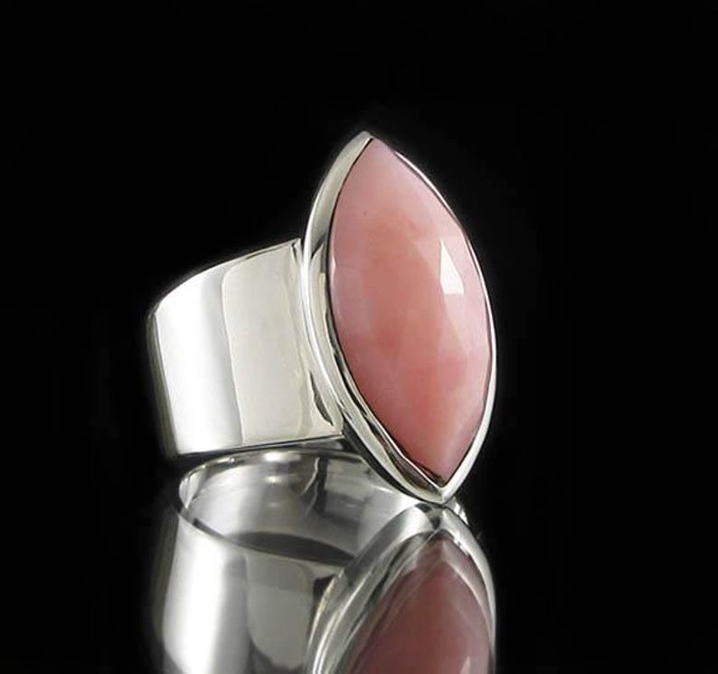 Pode incluir: Um anel de prata com uma grande pedra de opala rosa em forma de marquesa. O anel tem uma banda larga e um acabamento liso e polido.