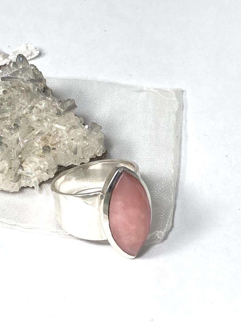 Pode incluir: Um anel de prata com uma gema rosa em forma de marquise. O anel est&aacute; sobre um fundo branco.