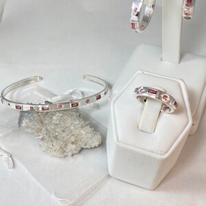 May include: A set of three silver jewelry pieces with pink gemstones. The set includes a ring, a bracelet, and a pair of earrings.