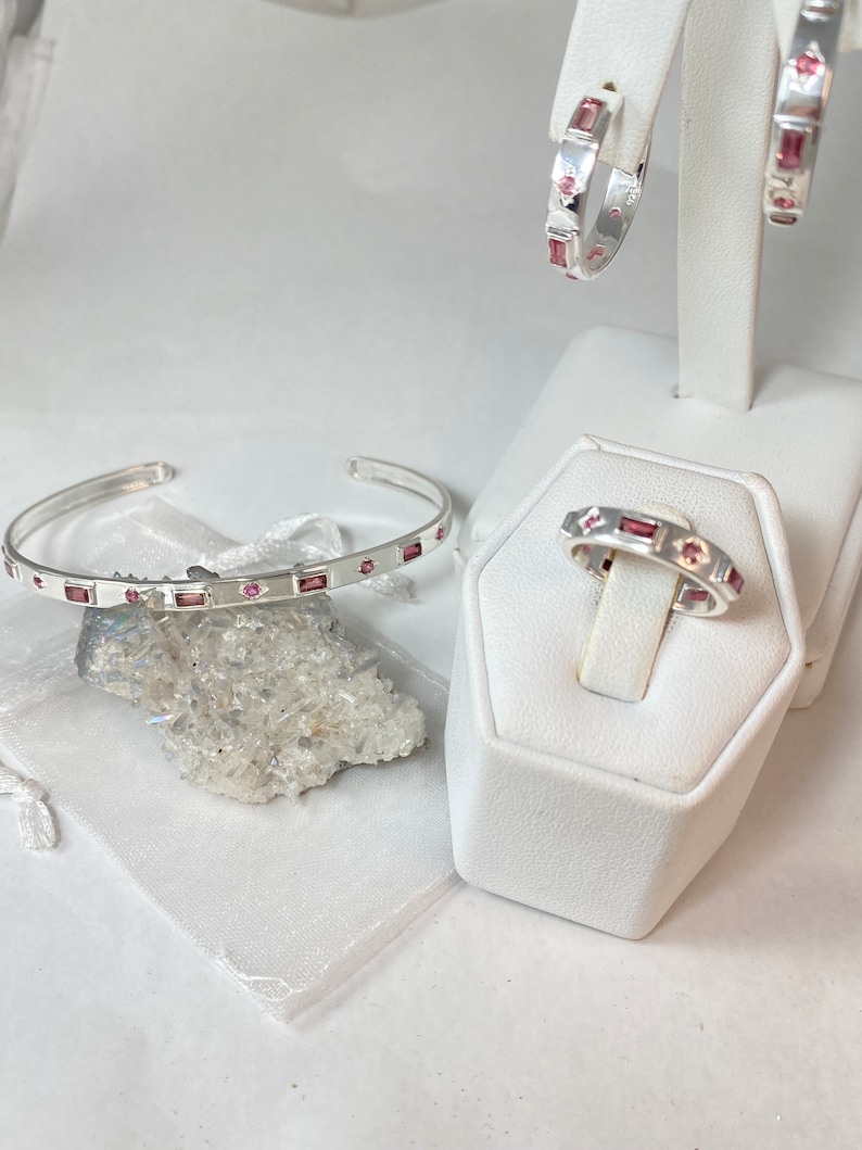 May include: A set of three silver jewelry pieces with pink gemstones. The set includes a ring, a bracelet, and a pair of earrings.