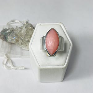 Pode incluir: Um anel de prata com uma gema rosa em forma de marquise engastada no centro. O anel &eacute; exibido em um suporte de anel branco.