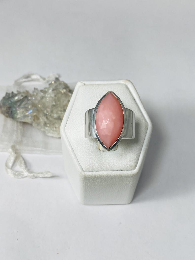 Pode incluir: Um anel de prata com uma gema rosa em forma de marquise engastada no centro. O anel &eacute; exibido em um suporte de anel branco.