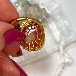 May include: A gold-toned ring featuring an oval, peach-colored gemstone. The ring's setting has a detailed, openwork design. The gemstone is held in place by small prongs. The ring is held by a person with pink nail polish.