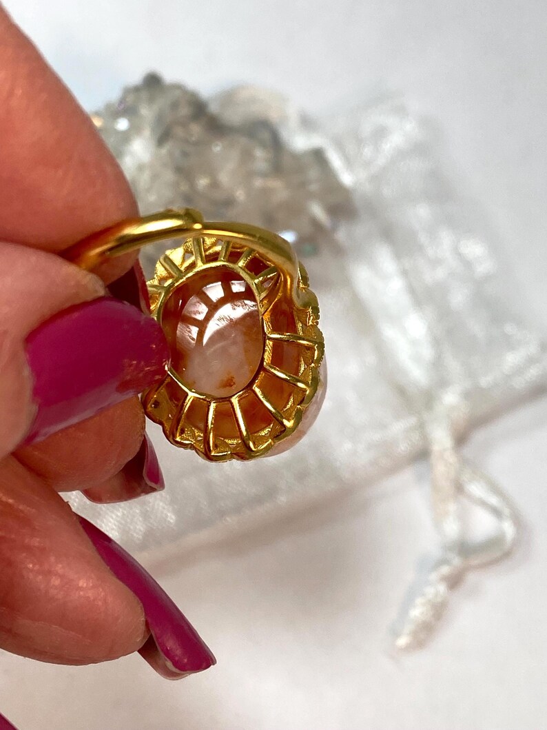 May include: A gold-toned ring featuring an oval, peach-colored gemstone. The ring's setting has a detailed, openwork design. The gemstone is held in place by small prongs. The ring is held by a person with pink nail polish.