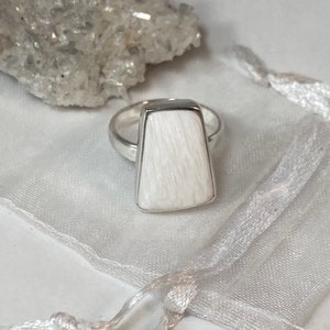 May include: A white rectangular ring with a silver band. The ring is made of a white stone with a textured surface.