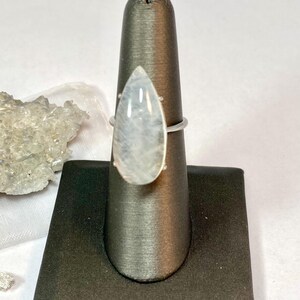 May include: A silver ring featuring a large, pear-shaped, iridescent moonstone. The stone is set in a simple, minimalist silver band. The ring is displayed on a black jewelry stand against a white background. The moonstone has a soft, milky appearance.