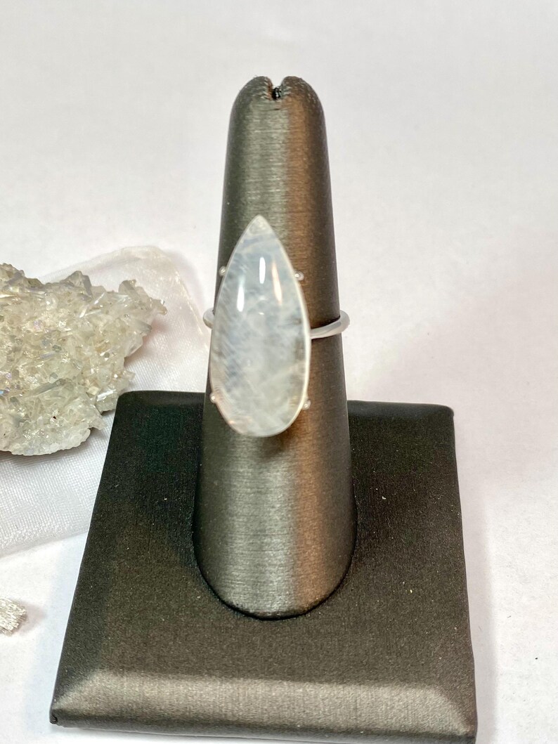 May include: A silver ring featuring a large, pear-shaped, iridescent moonstone. The stone is set in a simple, minimalist silver band. The ring is displayed on a black jewelry stand against a white background. The moonstone has a soft, milky appearance.