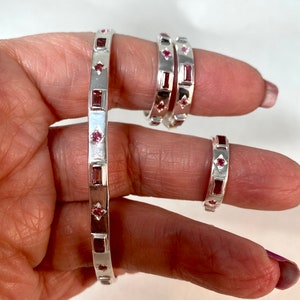 May include: Three silver rings with pink gemstones. The rings are stacked on a finger. The rings are all the same design, but different sizes.