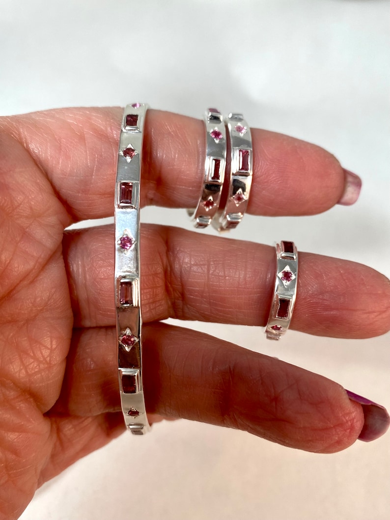 May include: Three silver rings with pink gemstones. The rings are stacked on a finger. The rings are all the same design, but different sizes.