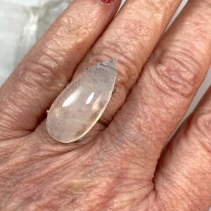 May include: A large, pear-shaped, clear gemstone ring with a silver band. The gemstone has a milky, translucent appearance. The ring is worn on a finger, with a red nail polish visible in the background. The ring is a statement piece.
