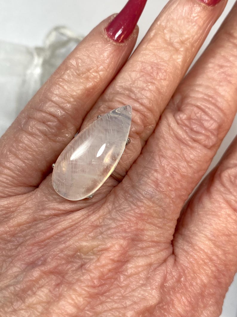 May include: A large, pear-shaped, clear gemstone ring with a silver band. The gemstone has a milky, translucent appearance. The ring is worn on a finger, with a red nail polish visible in the background. The ring is a statement piece.