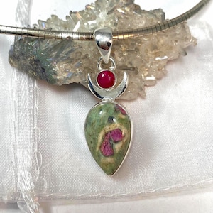May include: A silver pendant featuring a teardrop-shaped zoisite stone with embedded ruby inclusions, topped with a small round ruby cabochon set in a crescent moon frame, suspended from a silver omega chain necklace.