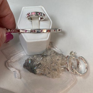 May include: A set of three silver jewelry pieces with pink gemstones. The set includes a ring, a bangle bracelet, and a pair of hoop earrings.