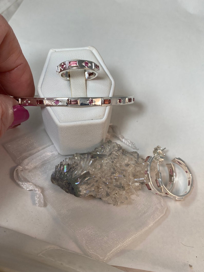 May include: A set of three silver jewelry pieces with pink gemstones. The set includes a ring, a bangle bracelet, and a pair of hoop earrings.