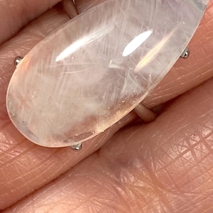 May include: A large, teardrop-shaped ring featuring a translucent, pale pink gemstone. The stone is set on a silver band with four small prongs. The ring is shown on a person's hand, highlighting the size and shape of the gemstone.