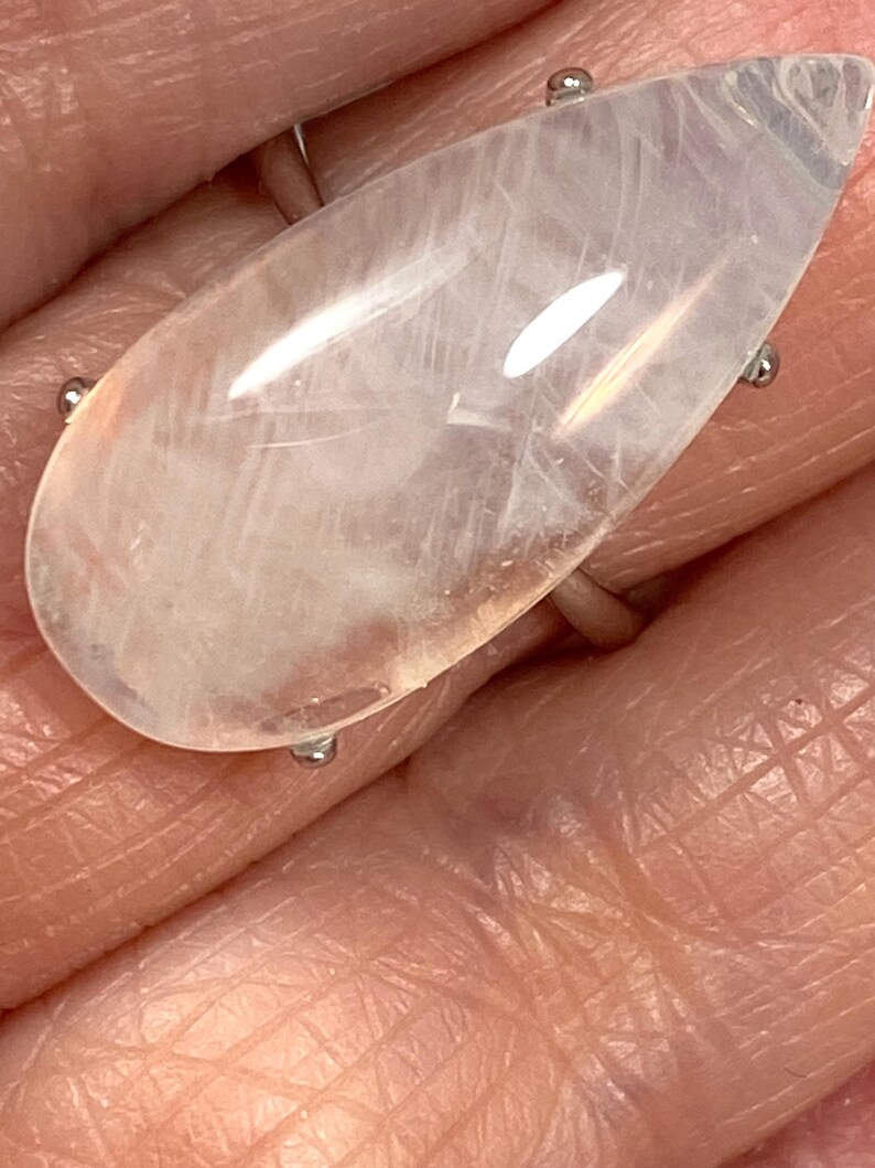 May include: A large, teardrop-shaped ring featuring a translucent, pale pink gemstone. The stone is set on a silver band with four small prongs. The ring is shown on a person's hand, highlighting the size and shape of the gemstone.