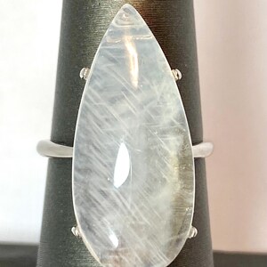 May include: A large, pear-shaped, clear gemstone ring with a silver band. The gemstone has internal striations and is mounted on a black display stand. The ring's design is simple yet elegant, suitable for everyday wear or special occasions.
