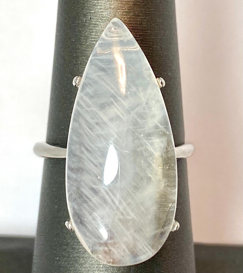 May include: A large, pear-shaped, clear gemstone ring with a silver band. The gemstone has internal striations and is mounted on a black display stand. The ring's design is simple yet elegant, suitable for everyday wear or special occasions.