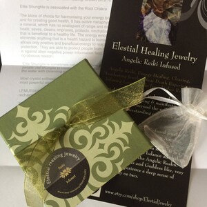May include: A green box with a gold ribbon and a white label that says "Elestial Healing Jewelry Reiki Infused" with a website address. The box is sitting on a black card with white text that says "Elestial Healing Jewelry Angelic Reiki Infused" and includes information about the benefits of the jewelry.