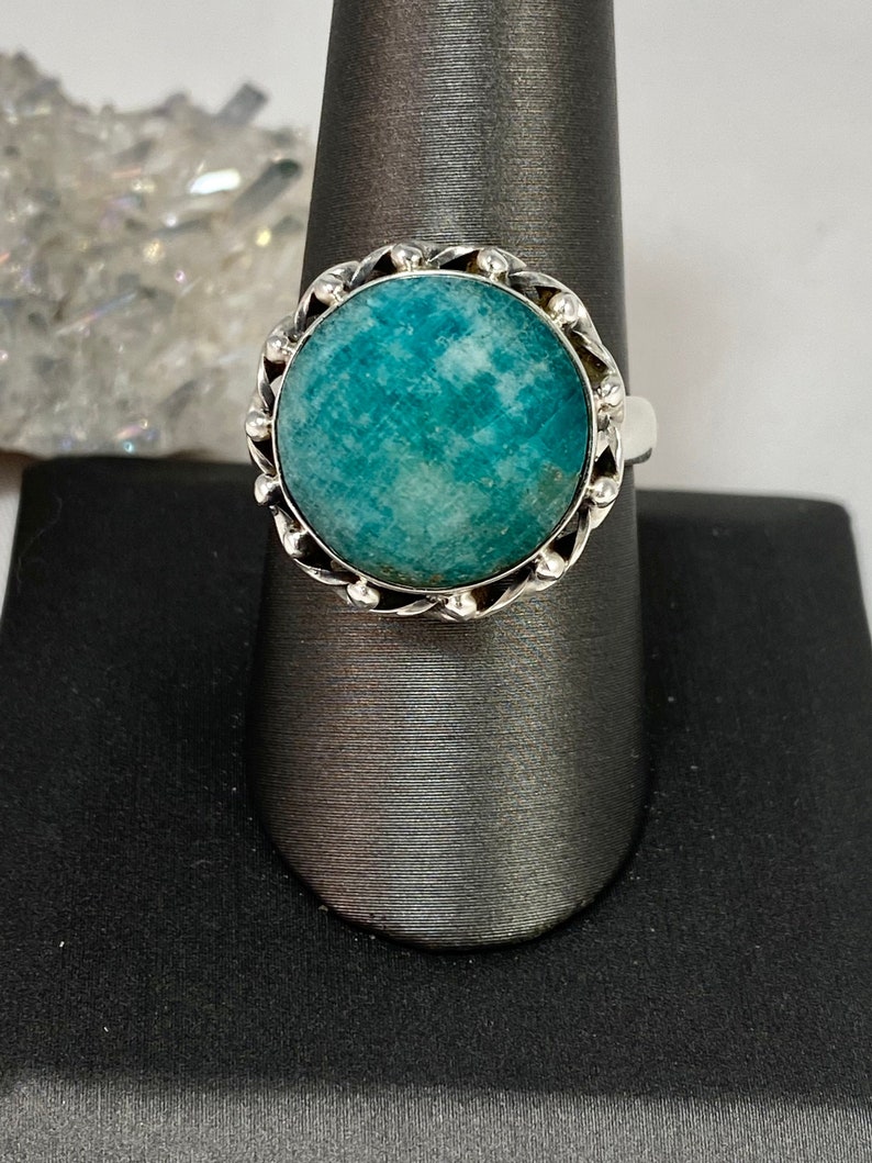 K&ouml;nnte beinhalten: Ein silberner Ring mit einem gr&uuml;nen Edelstein, der in einer dekorativen Fassung sitzt. Der Ring befindet sich auf einem grauen Pr&auml;sentationsst&auml;nder.