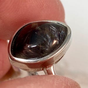 May include: A silver ring featuring a dark, oval-shaped gemstone with internal, hair-like inclusions. The gemstone has a polished, reflective surface, and the silver band is smooth and shiny. The ring is held up by a finger.