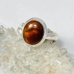 May include: A silver ring with a large oval-shaped brown gemstone. The gemstone has a reddish-brown color and a shiny, polished finish.