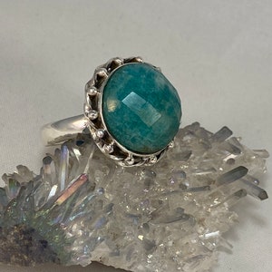 K&ouml;nnte beinhalten: Ein silberner Ring mit einem gro&szlig;en, facettierten, blaugr&uuml;nen Edelstein, der in einer dekorativen, perlenbesetzten Kronenfassung sitzt.