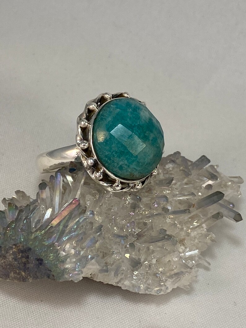 K&ouml;nnte beinhalten: Ein silberner Ring mit einem gro&szlig;en, facettierten, blaugr&uuml;nen Edelstein, der in einer dekorativen, perlenbesetzten Kronenfassung sitzt.