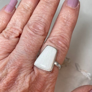 May include: A white, rectangular ring with a silver band. The ring is made of a white, striated material.