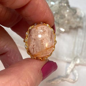 May include: A gold ring with a large, oval, peach-colored stone. The stone has a marbled appearance with light brown and white patterns. The ring's band is gold with a decorative, diamond-like border around the stone.