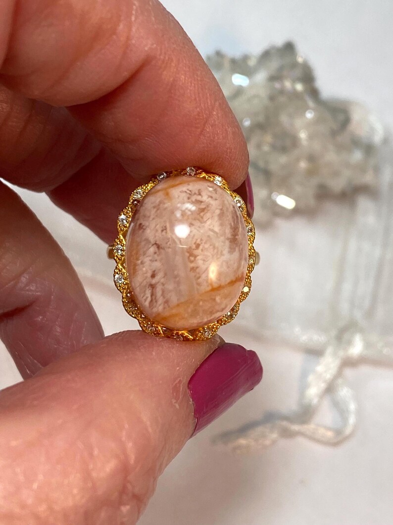 May include: A gold ring with a large, oval, peach-colored stone. The stone has a marbled appearance with light brown and white patterns. The ring's band is gold with a decorative, diamond-like border around the stone.