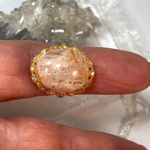 May include: A gold-toned ring featuring a large, oval-shaped gemstone with a marbled appearance in shades of beige and brown. The gemstone is encircled by a delicate gold setting adorned with small, clear crystals, creating a halo effect. The ring is a piece of jewelry.