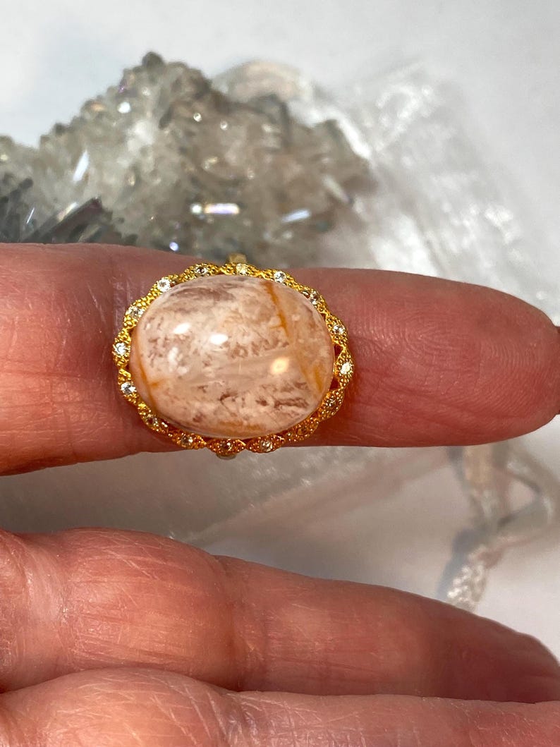 May include: A gold-toned ring featuring a large, oval-shaped gemstone with a marbled appearance in shades of beige and brown. The gemstone is encircled by a delicate gold setting adorned with small, clear crystals, creating a halo effect. The ring is a piece of jewelry.