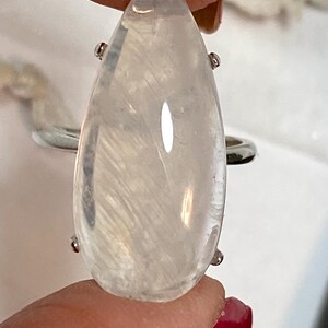 May include: A clear, teardrop-shaped gemstone ring with a silver band. The gemstone has internal white feather-like inclusions and is held in place by four small silver prongs. The ring is a piece of jewelry.