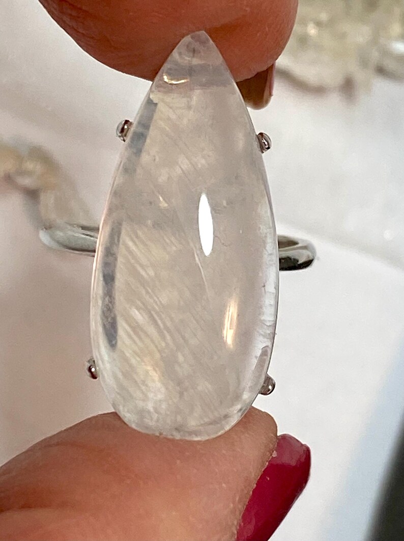 May include: A clear, teardrop-shaped gemstone ring with a silver band. The gemstone has internal white feather-like inclusions and is held in place by four small silver prongs. The ring is a piece of jewelry.