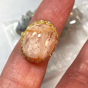 May include: A gold ring featuring an oval, peach-colored gemstone. The gemstone is surrounded by a gold filigree setting with small, clear accent stones. The ring is displayed on a finger, showcasing its design and color.
