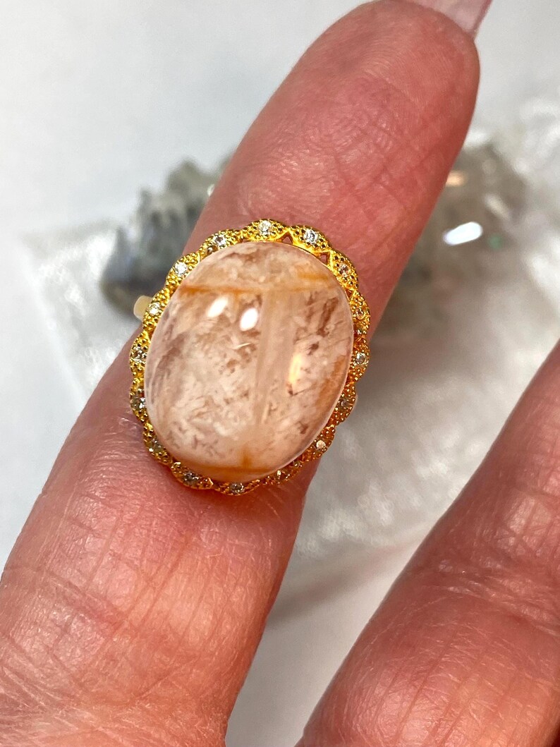 May include: A gold ring featuring an oval, peach-colored gemstone. The gemstone is surrounded by a gold filigree setting with small, clear accent stones. The ring is displayed on a finger, showcasing its design and color.