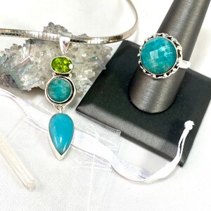 K&ouml;nnte beinhalten: Eine silberne Halskette mit einem tropfenf&ouml;rmigen blauen Edelstein-Anh&auml;nger und einem kleineren runden blauen Edelstein und einem gr&uuml;nen Edelstein dar&uuml;ber. Ein silberner Ring mit einem runden blauen Edelstein ist ebenfalls abgebildet.