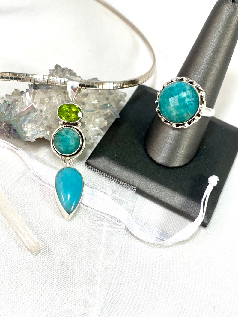 K&ouml;nnte beinhalten: Eine silberne Halskette mit einem tropfenf&ouml;rmigen blauen Edelstein-Anh&auml;nger und einem kleineren runden blauen Edelstein und einem gr&uuml;nen Edelstein dar&uuml;ber. Ein silberner Ring mit einem runden blauen Edelstein ist ebenfalls abgebildet.