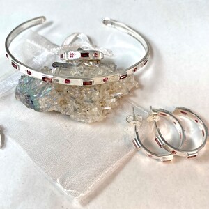 May include: A set of silver jewelry with pink gemstones. The set includes a bangle bracelet, a ring, and a pair of hoop earrings.